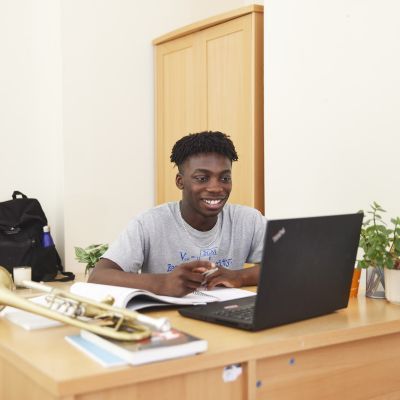 A boy is sat at a desk, smiling at his laptop